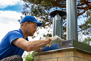 Chimney service Freehold professional in a blue shirt and cap wearing work gloves working on a chimney cap.